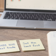 Why a big win can wreck you worse than a loss Calm wooden desk with three yellow sticky notes reading "Win = data, not proof", "Same size next trade" and "Set the limit first", beside a notebook, blue Bic pen, coffee and a laptop showing a candlestick chart.