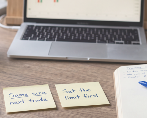 Calm wooden desk with three yellow sticky notes reading "Win = data, not proof", "Same size next trade" and "Set the limit first", beside a notebook, blue Bic pen, coffee and a laptop showing a candlestick chart.