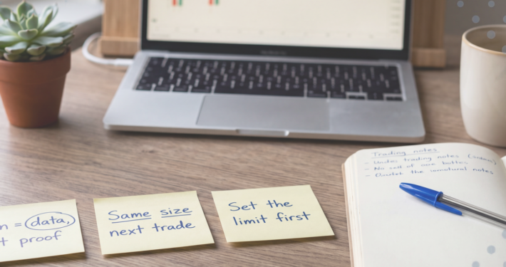 Calm wooden desk with three yellow sticky notes reading "Win = data, not proof", "Same size next trade" and "Set the limit first", beside a notebook, blue Bic pen, coffee and a laptop showing a candlestick chart.