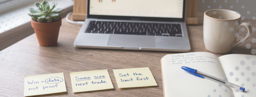 Calm wooden desk with three yellow sticky notes reading "Win = data, not proof", "Same size next trade" and "Set the limit first", beside a notebook, blue Bic pen, coffee and a laptop showing a candlestick chart.