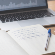 Why you freeze on a valid setup but click instantly on a bad one A calm wooden trading desk with an open notebook, a blue Bic pen and a defocused candlestick chart on a laptop, beside two yellow sticky notes reading WAIT and CLICK.