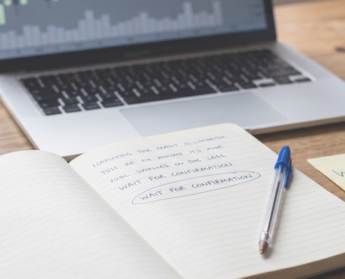 A calm wooden trading desk with an open notebook, a blue Bic pen and a defocused candlestick chart on a laptop, beside two yellow sticky notes reading WAIT and CLICK.