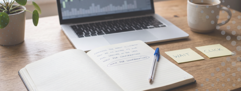 A calm wooden trading desk with an open notebook, a blue Bic pen and a defocused candlestick chart on a laptop, beside two yellow sticky notes reading WAIT and CLICK.