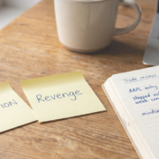 Revenge trading dressed up as conviction — hero Two yellow sticky notes on a warm wooden desk labelled CONVICTION and REVENGE, beside an open trading journal with handwritten notes and a blue Bic pen, laptop with candlestick chart in background, natural daylight.