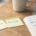 Two yellow sticky notes on a warm wooden desk labelled CONVICTION and REVENGE, beside an open trading journal with handwritten notes and a blue Bic pen, laptop with candlestick chart in background, natural daylight.