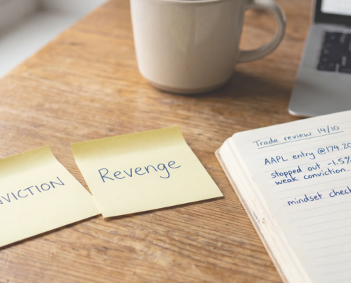 Two yellow sticky notes on a warm wooden desk labelled CONVICTION and REVENGE, beside an open trading journal with handwritten notes and a blue Bic pen, laptop with candlestick chart in background, natural daylight.