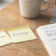 Two yellow sticky notes on a warm wooden desk labelled CONVICTION and REVENGE, beside an open trading journal with handwritten notes and a blue Bic pen, laptop with candlestick chart in background, natural daylight.