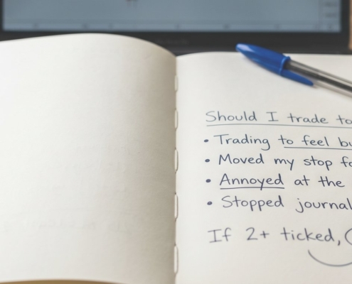 A flat-bound notebook open on a wooden desk with a handwritten pre-session check headed 'Should I trade today?', four warning-sign questions, and 'flat day' circled, a blue Bic pen on the page.