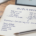 Open notebook on a wooden desk with dark blue handwritten notes headed Win rate vs expectancy, comparing a 70% and a 40% win rate, an expectancy formula and a Track instead list, beside a blue Bic pen and a coffee mug, with a calm candlestick chart on a laptop behind.