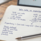 Open notebook on a wooden desk with dark blue handwritten notes headed Win rate vs expectancy, comparing a 70% and a 40% win rate, an expectancy formula and a Track instead list, beside a blue Bic pen and a coffee mug, with a calm candlestick chart on a laptop behind.
