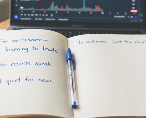 A trading journal reading I'm learning to trade beside a laptop chart on a wooden desk