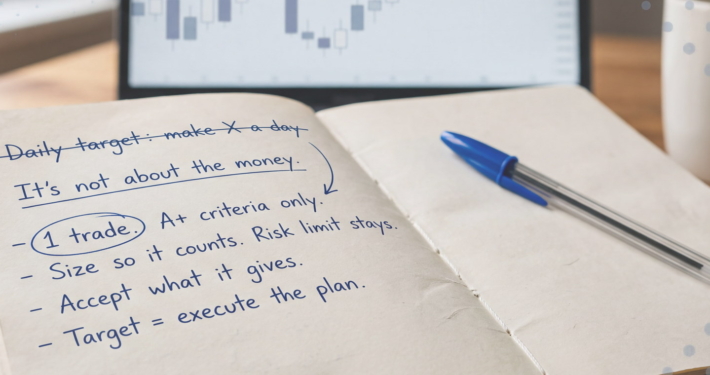 Open notebook on a wooden desk with handwritten notes in blue ink: "Daily target: make X a day" crossed out, then "It's not about the money" and a list starting with a circled "1 trade".